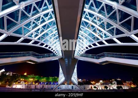 Brücke des Friedens in der Nacht in Tiflis, Georgien. Moderne Fußgängerbrücke über den Fluss Kura. Symmetrisches Design mit Treppen. Stockfoto