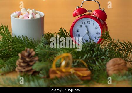 Weihnachtsschmuck und rote Uhr auf dem Tisch mit Pine Cone, Zimt, Nüssen und Tasse Kaffee mit Marshmallows Stockfoto