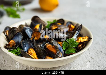 Köstliche frisch gedünstete Muscheln in Weißweinsauce auf einem weißen Teller, eine Zitrone und Petersilie auf grauem Grund Stockfoto