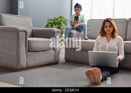 EIN SIKH-JUNGE SPIELT MIT DEM HANDY, WÄHREND ER AUF DEM SOFA SITZT MIT MUTTER, DIE AN EINEM LAPTOP AUF DEM BODEN SITZT Stockfoto