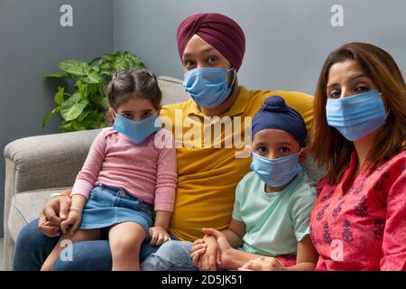 EINE SIKH FAMILIE SITZT ZUSAMMEN AUF DEM SOFA MIT MASKEN UND BLICK AUF DIE KAMERA Stockfoto