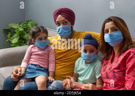 EINE SIKH FAMILIE SITZT ZUSAMMEN AUF DEM SOFA MIT MASKEN UND BLICK AUF DIE KAMERA Stockfoto