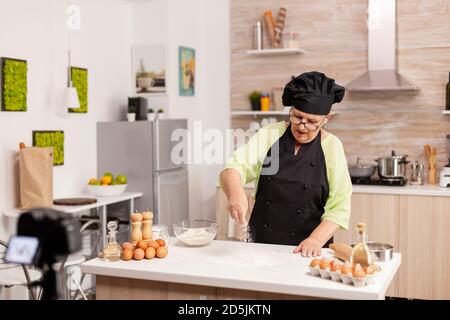 Ältere Frau Erstellen von Inhalten für kulinarische Blog Zubereitung von leckeren Brot. Pensionierter Blogger Chef Influencer mit Internet-Technologie kommunizieren, schießen Blogging auf Social Media mit digitalen Geräten. Stockfoto