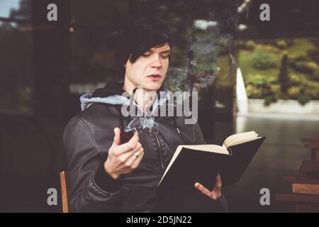 Junger schwarzhaariger Mann, der im Café im Freien ein Buch durchbohrt und Tabakpfeife raucht. Stockfoto