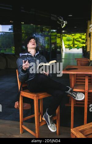 Junger schwarzhaariger Mann, der im Café im Freien ein Buch durchbohrt und Tabakpfeife raucht. Stockfoto