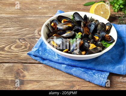 Köstliche frisch gedünstete Muscheln in Weißweinsauce auf einem weißen Teller, eine Zitrone und Petersilie auf einem Holztisch Stockfoto