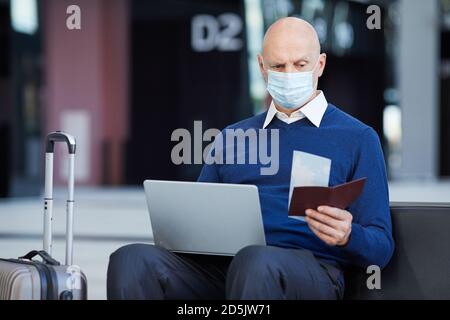 Reifer Mann in Maske mit Laptop er den Austausch der Tickets Sitzen am Flughafen Stockfoto