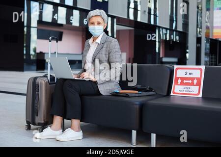 Reife Frau in Schutzmaske Blick auf Kamera während des Sitzens Im Warteraum während der Pandemie und mit Laptop-Computer Stockfoto