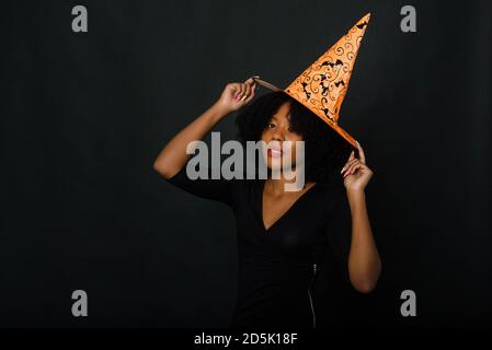 Horizontale Aufnahme einer fröhlichen jungen schwarzen Frau mit Afro-Haarschnitt, die einen orangen Halloween-Hut und ein schwarzes Kleid trägt. Studio Porträt auf schwarzem Hintergrund. Stockfoto