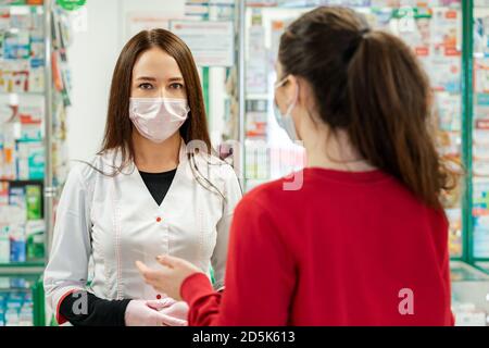 Eine Apothekerin in einer medizinischen Maske und Handschuhen bedient einen Kunden. Blick von der Schulter des Kunden. Das Konzept der Kauf von Medikamenten in einem Pharm Stockfoto