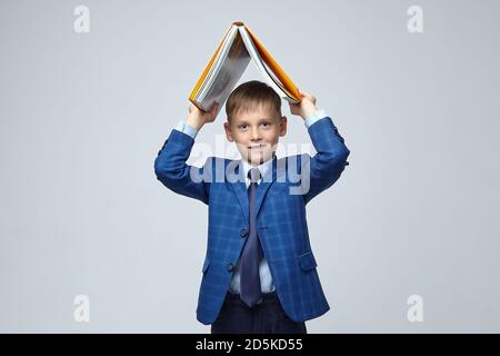 Schöner blonder Junge in einem Schulanzug, hält ein Buch über den Kopf mit einem Lächeln Stockfoto