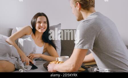 Schuss von einem fürsorglichen jungen Mann bringt Tablett mit Frühstück im Bett zu seinem schönen weiblichen Partner. Ideal Tender Beziehung und Kaffee und Croissants in Stockfoto