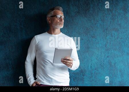 Serious Casual Grey-haired Reife männliche Führungskraft mit digitalen Tablet vor dunkelblauem Hintergrund. Stockfoto