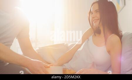 Schuss von einem fürsorglichen jungen Mann bringt Tablett mit Frühstück im Bett zu seinem schönen weiblichen Partner. Ideal Tender Beziehung und Kaffee und Croissants in Stockfoto