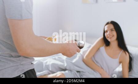 Schuss von einem fürsorglichen jungen Mann bringt Tablett mit Frühstück im Bett zu seinem schönen weiblichen Partner. Ideal Tender Beziehung und Kaffee und Croissants in Stockfoto