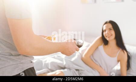 Schuss von einem fürsorglichen jungen Mann bringt Tablett mit Frühstück im Bett zu seinem schönen weiblichen Partner. Ideal Tender Beziehung und Kaffee und Croissants in Stockfoto