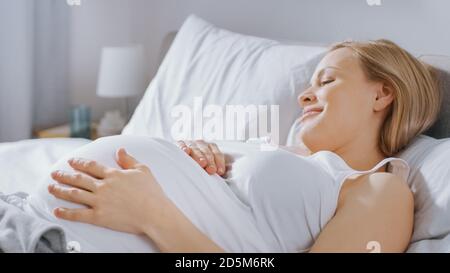 Glückliche junge schwangere Frau, die morgens im Bett liegt, berührt und streichelt sie ihren Bauch zärtlich, liebt ihr zukünftiges Baby und jeden Moment von ihr Stockfoto