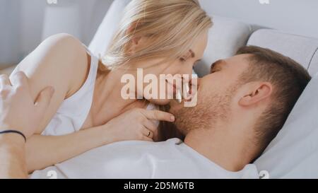 Happy Young couple Kuscheln zusammen im Bett, küssen und lächeln. Schöne Blonde weiblich und sie fit liebevolle Partner erleben Ausschreibung und Stockfoto