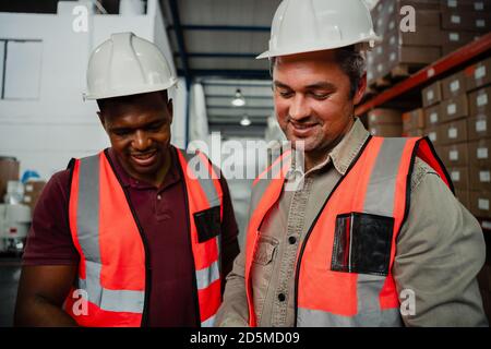 Lächelnde Kollegen tragen Overalls und Hüte, die in der Fabrik lachen Stockfoto