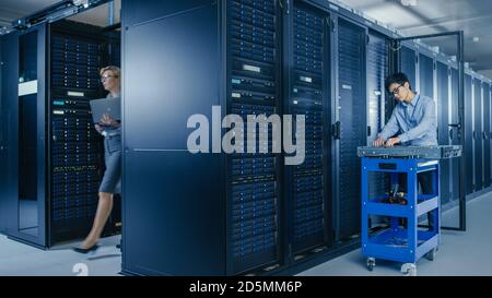 Im modernen Rechenzentrum: Team von IT-Technikern, die mit Server-Racks arbeiten, Mann mit Pushcart wechselt fehlerhafte Festplatten, Hardware-Wartung Stockfoto