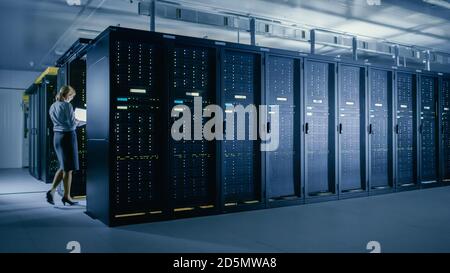 Im Rechenzentrum: Weibliche IT-Technikerin geht zwischen Reihen von Server-Racks, verwendet Laptop-Computer, um Wartungsdiagnose auszuführen, damit Mainframe funktioniert Stockfoto