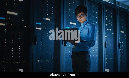 In Dark Data Center: Männlicher IT-Spezialist geht entlang der Reihe der operativen Server-Racks, nutzt Laptop für die Wartung. Konzept für Cloud Computing Stockfoto