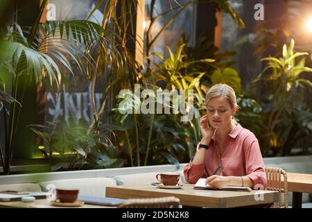 Junge Frau, die am Tisch sitzt und Notizen macht Notieren Sie sich das Pad, während Sie Kaffee im Café trinken Stockfoto