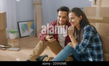 Happy Young Paar sitzt auf dem Boden der neu Vermietet / gekauft Wohnung und nutzen Smartphone. Unverpackte Kartons und bedeckte Möbel Stockfoto
