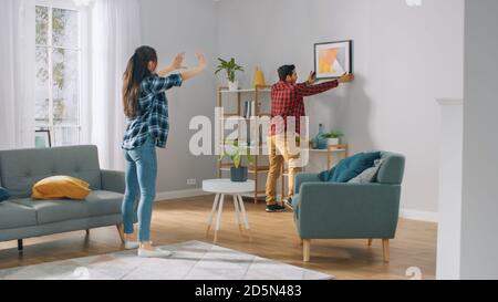 Happy couple Hanging Bild an der Wand, Freund bewegt es, Freundin sagt ihm, wenn der Rahmen ist gerade hängend. Lustige Moment in jungen Paares Stockfoto