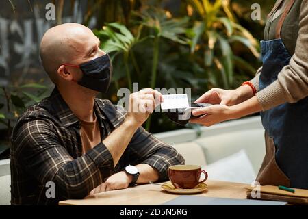 Junger Mann in Schutzmaske für seine Bestellung mit bezahlen Kreditkarte im Café Stockfoto