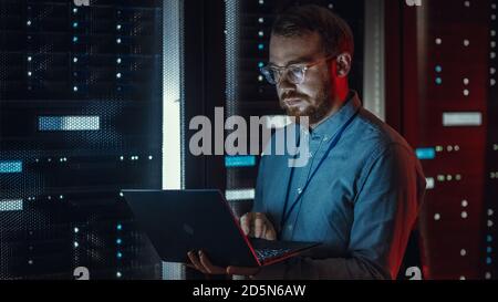 Bared IT Specialist in Glasses arbeitet an Laptops im Rechenzentrum, während er in der Nähe des Server-Racks steht. Diagnose Ausführen, Wartungsarbeiten Durchführen Stockfoto