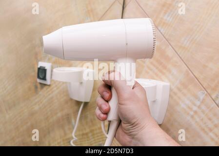 Weißer Haartrockner in Nahaufnahme. Ein Mann hält einen Plastiktrockner in der Hand vor einem Hintergrund aus Spiegeln und Marmorfliesen. Interieur eines Badezimmers, Schönheit Stockfoto