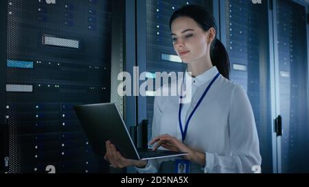 Selbstbewusste und schöne IT-Ingenieurin arbeitet im Rechenzentrum an einem Laptop, während sie vor dem Server Rack steht. Diagnose wird ausgeführt oder ausgeführt Stockfoto