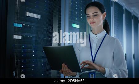 Selbstbewusste und schöne IT-Ingenieurin arbeitet im Rechenzentrum an einem Laptop, während sie vor dem Server Rack steht. Diagnose wird ausgeführt oder ausgeführt Stockfoto