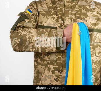 Ukrainischer Mann-Krieger, der mit einer Militär-Pixel-Uniform bekleidet ist, hält die gelb-blaue Flagge des Staates Ukraine und an der Kette ein kleines Wappen von Stockfoto