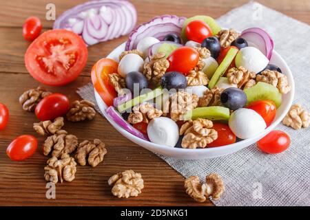 Salat mit frischen Kirschtomaten, Mozzarella-Käse, Oliven, Kiwi, Zwiebel und Walnüssen auf braunem Holzhintergrund, Kopierraum, Nahaufnahme. Stockfoto