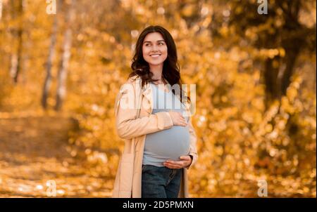 Schöne junge schwangere Frau umarmt ihren Bauch auf zu Fuß in Herbstpark Stockfoto