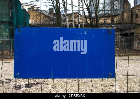 Leeres blaues Schild, das einen Zaun überreicht. Schild am Drahtzaun. Altes Gebäude im Hintergrund. Mit Kopierbereich signieren. Blaues Rechteck. Straßenschild. Stockfoto