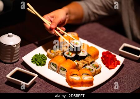 Person, die Sushi mit Essstäbchen im Restaurant isst. Crop anonyme Person mit Essstäbchen essen verschiedene Arten von Sushi-Rollen serviert mit Wasabi und Stockfoto
