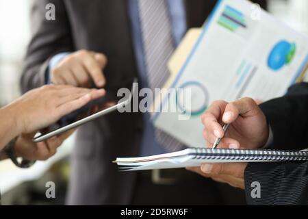 Mann im Geschäftsanzug mit Papieren in den Händen schreibt mit Kugelschreiber mit Kollegen auf Hintergrund in Büro Nahaufnahme Stockfoto