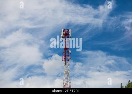 Ein Zellenturm aus rotem und weißem Metall gegen Ein blauer Himmel mit weißen Wolken in den Altai-Bergen Vor einem Hintergrund von grünen Baumkronen für breit Stockfoto