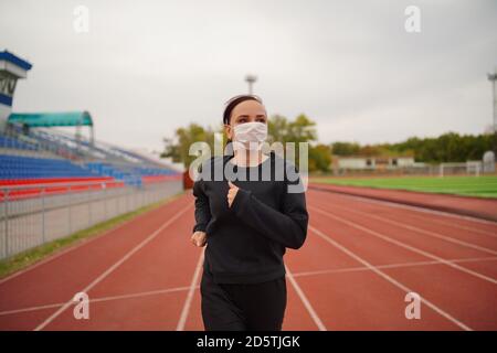Aktive Frau in Schutzmaske läuft im Stadion. Young fit female in Sportbekleidung und Schutzmaske für Coronavirus Prävention laufen auf roter Spur Stockfoto