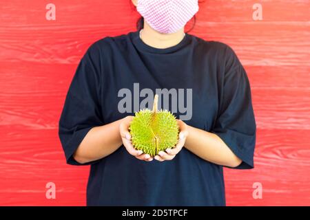 Junge Frau trägt eine Schutzmaske, um eine Infektion des Corona-Virus (Covid-19) in einem schwarzen Hemd mit einem reifen Durian zu verhindern, bereit, mit dem roten w zu essen Stockfoto