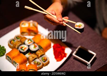 Person, die Sushi mit Essstäbchen im Restaurant isst. Crop anonyme Person mit Essstäbchen essen verschiedene Arten von Sushi-Rollen serviert mit Wasabi und Stockfoto