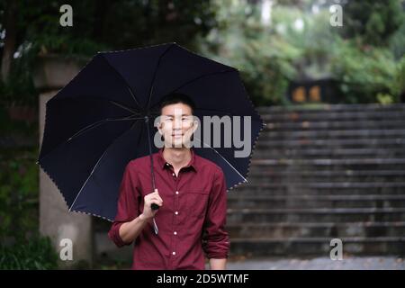 Mittellange Aufnahme eines jungen asiatischen Mannes, der unter einem Regenschirm und einem Lächeln auf die Kamera schaut. Im Naturpark Stockfoto