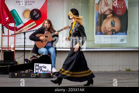 Dundee, Tayside, Schottland, Großbritannien. Oktober 2020. UK Wetter: Ein heller Tag mit gelegentlichem Sonnenschein und leichtem Wind über den Nordosten Schottlands, Höchsttemperaturen 13 Grad Eine sehr modische blonde Frau, die eine Gesichtsmaske und Haare trägt, die im leichten Wind wehen, kam während der Covid-19 Lockdown an einer Buskerin (Maria McAveety) vor der Boots-Apotheke im Stadtzentrum von Dundee vorbei. Kredit: Dundee Photographics/Alamy Live Nachrichten Stockfoto