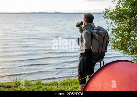 Mann, der in Estland campt, am See mit Rucksack steht und Kaffee trinkt Stockfoto