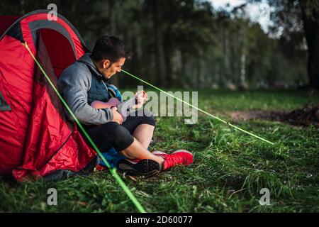 Mann, der in Estland zeltet, in seinem Zelt sitzt und die Ukulele spielt Stockfoto