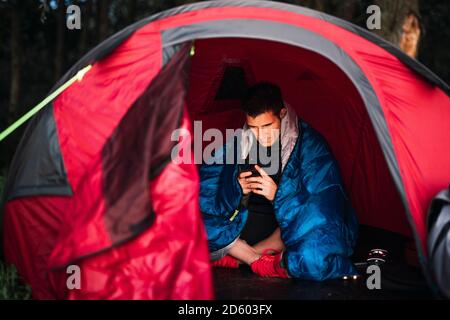 Mann, der in Estland campte, in seinem Zelt sitzend, mit dem Smartphone Stockfoto