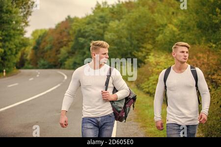 Sommerurlaub. Automatischer Stopp für Reisende. Budget Reisende. Männer Rucksäcke zu Fuß zusammen auf der Straße. Zwillinge zu Fuß entlang der Straße. Abenteuer und Entdeckung. Mann mit Rucksack Anhalter auf der Straße. Stockfoto